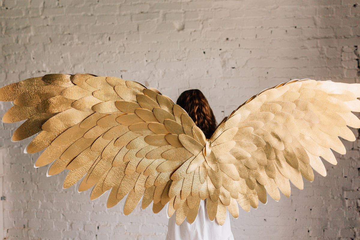 May include: A pair of large, golden angel wings made of feathers. The wings are spread wide and are in front of a white brick wall.