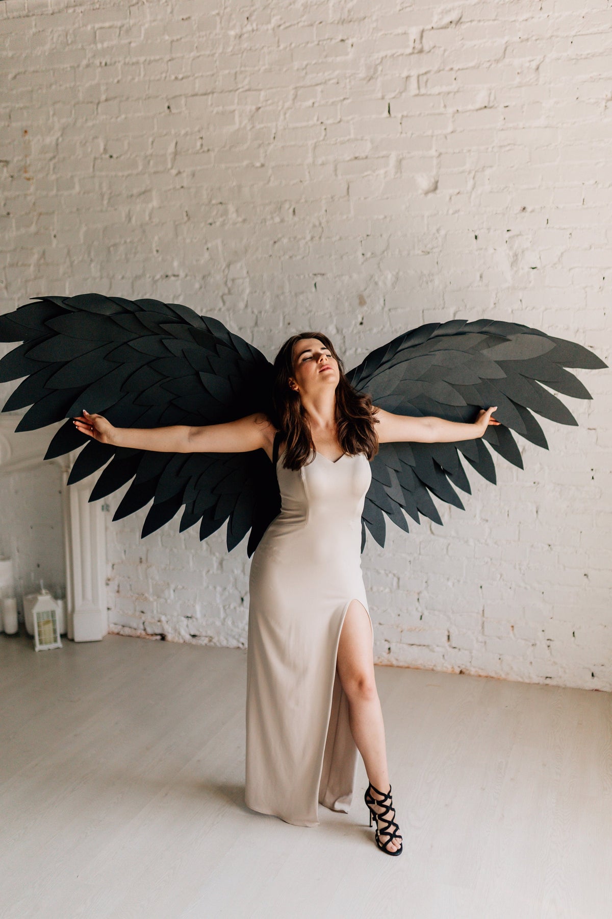 May include: A woman wearing a beige, floor-length dress with a high slit and black strappy heels, posing with large, black paper angel wings against a white brick wall.