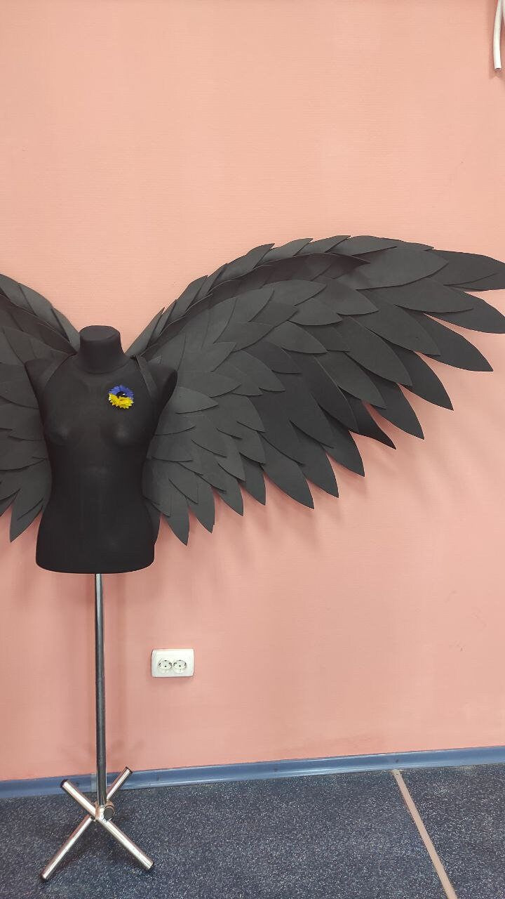 May include: A black mannequin torso with large, layered black wings. A small blue and yellow flower pin is attached to the mannequin. The wings are made of many individual, dark-colored pieces, creating a feathered effect. The background is a solid pink wall.