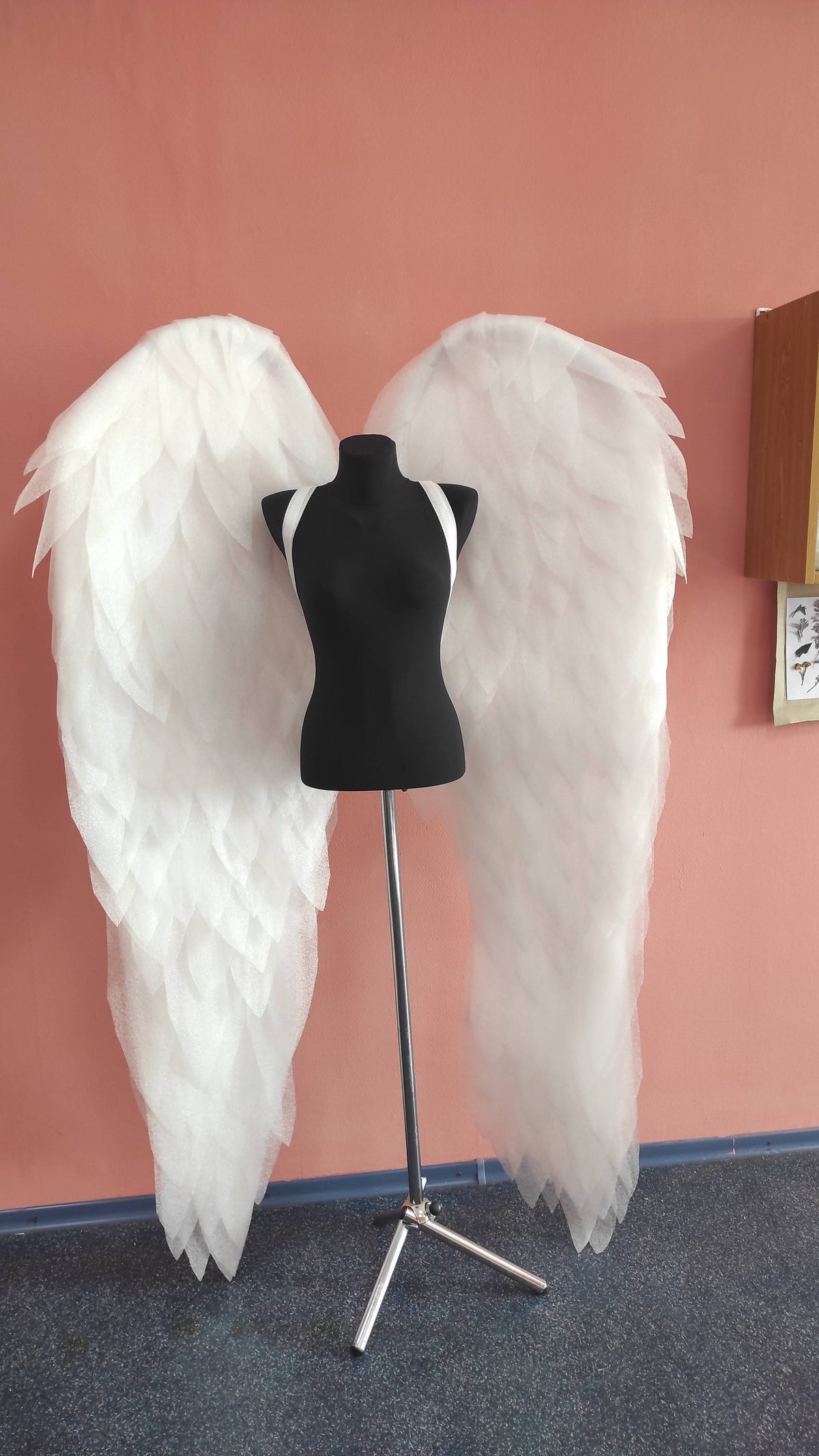 May include: A pair of large, white, feathery angel wings on a black mannequin stand against a pink wall.