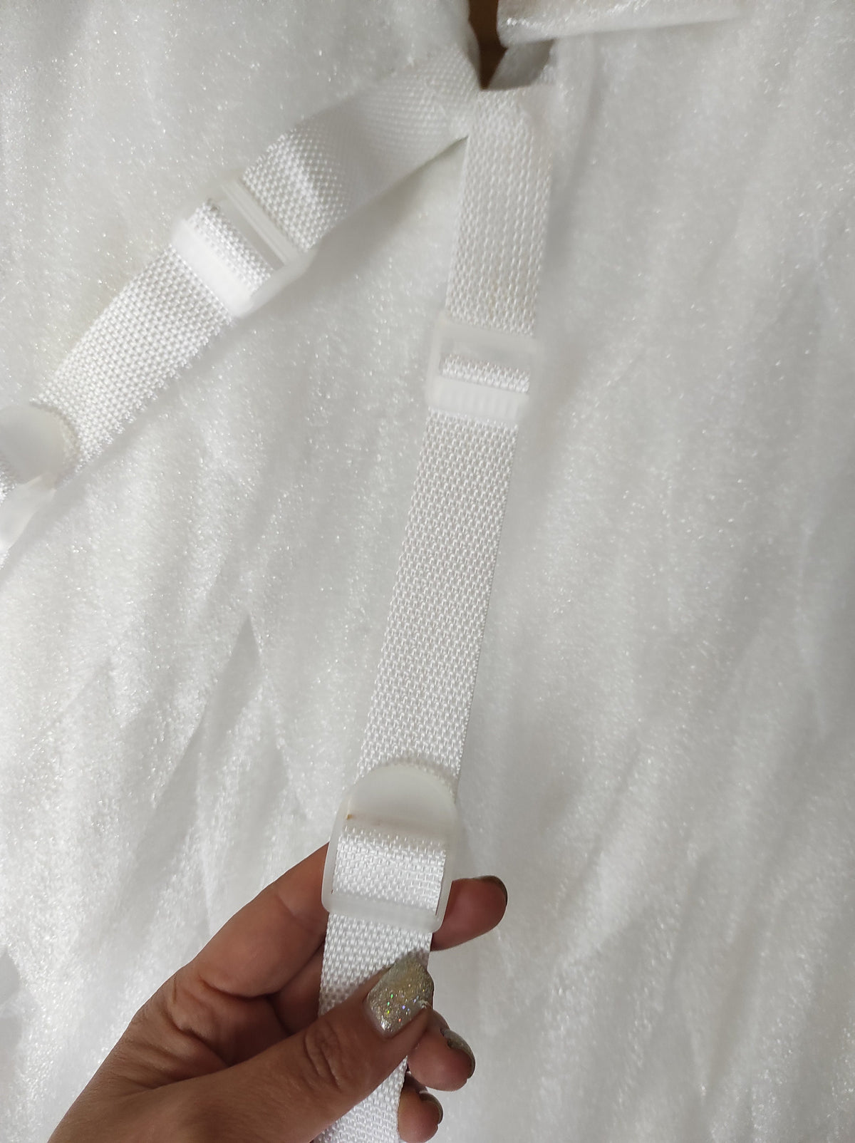 May include: A white woven strap featuring a plastic adjustable slider buckle. The strap is attached to a white textured material, and the buckle allows for length modification of the webbing. The design is functional and monochromatic in white.