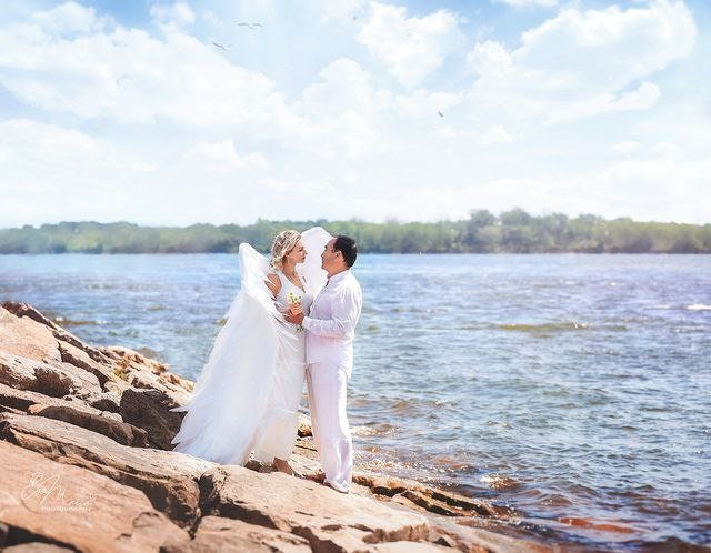 May include: A bride and groom in white clothing stand on a rocky shore by a body of water. The bride is wearing a white dress with a large white feathered wing attached to her back. The groom is wearing a white button-down shirt and white pants. The couple is looking at each other and smiling.