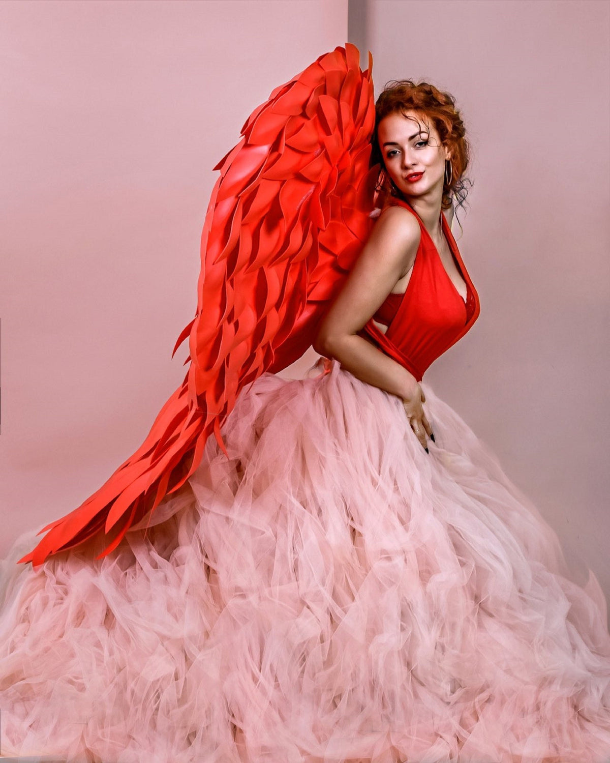May include: A woman wearing a red dress and a large, vibrant red wing costume. The dress has a plunging neckline and a full, layered pink skirt. The background is a soft pink.