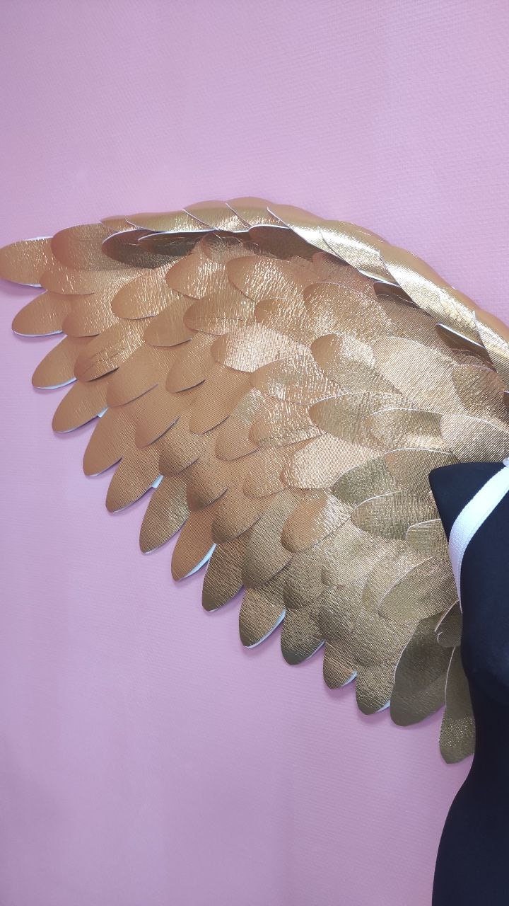 May include: A pair of large, gold angel wings with a textured, metallic finish. The wings are made of individual feathers that are layered and overlapping.