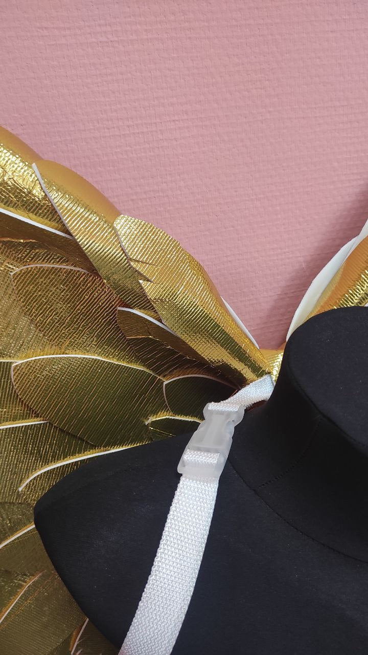 May include: Gold-colored costume wings with a textured, feather-like design. The wings are attached to a white strap with a clear plastic buckle. The wings are displayed on a black mannequin.