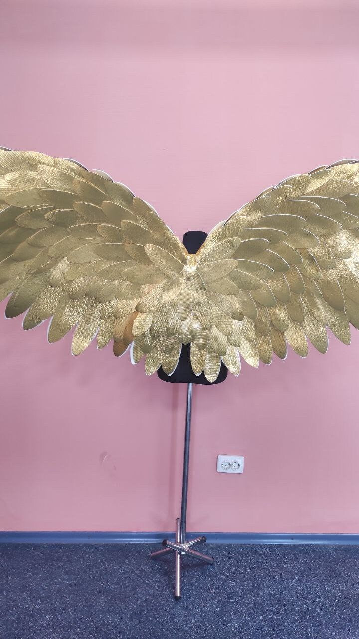 May include: Gold-colored angel wings with a layered feather design. The wings are displayed on a black mannequin form against a pink wall. The wings are made of a textured material.