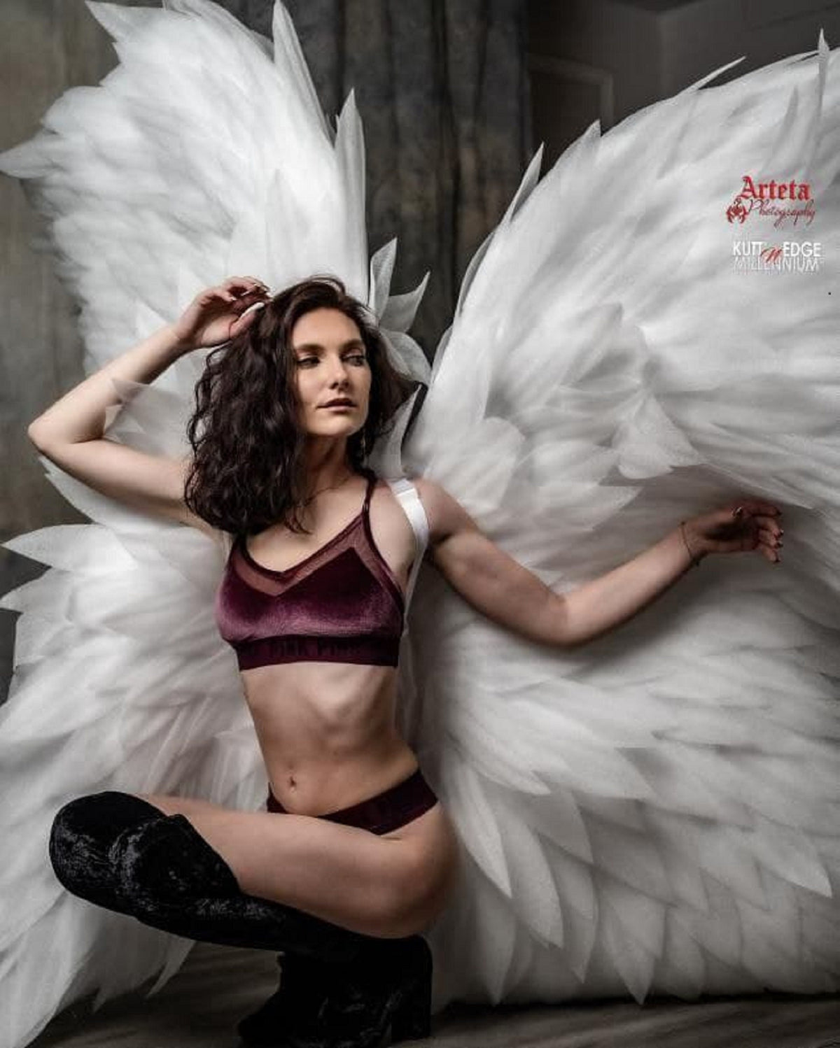 May include: A woman wearing a burgundy velvet bra and matching underwear poses with large white angel wings. The wings are made of feathers and are very detailed. The woman has long dark hair and is looking at the camera.