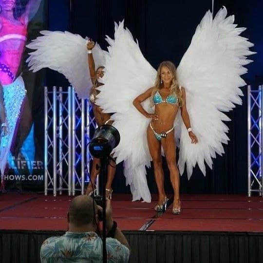 May include: Two women wearing white angel wings and blue and silver bikinis pose on a stage. One woman is holding a trophy. The stage is decorated with a red carpet and a silver backdrop.