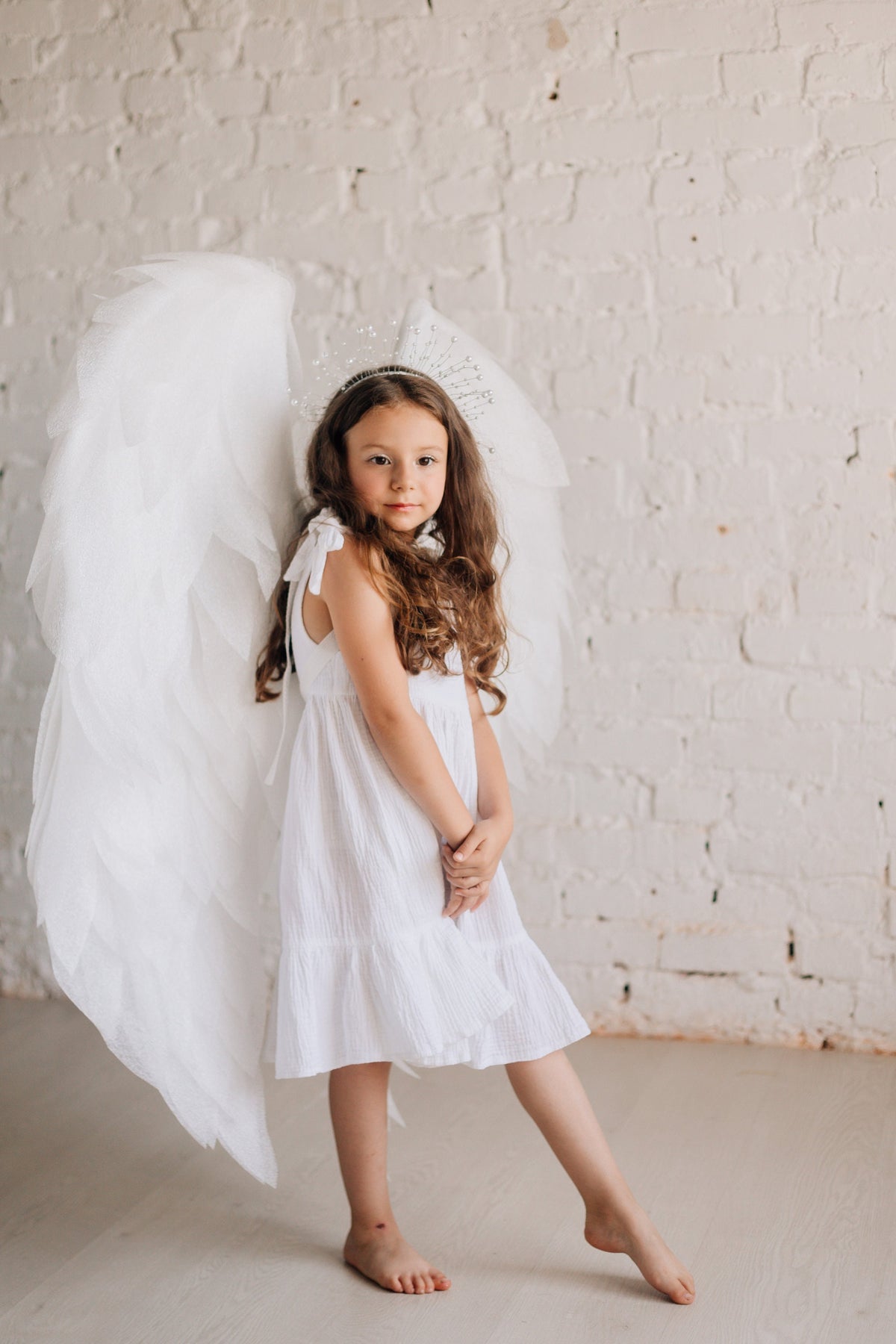 May include: A young girl wearing a white dress and a silver crown with white angel wings. She is standing in front of a white brick wall.