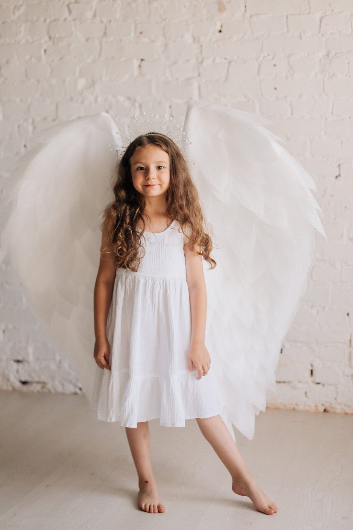 May include: A young girl wearing a white dress and angel wings with a silver crown on her head. She is standing in front of a white brick wall.