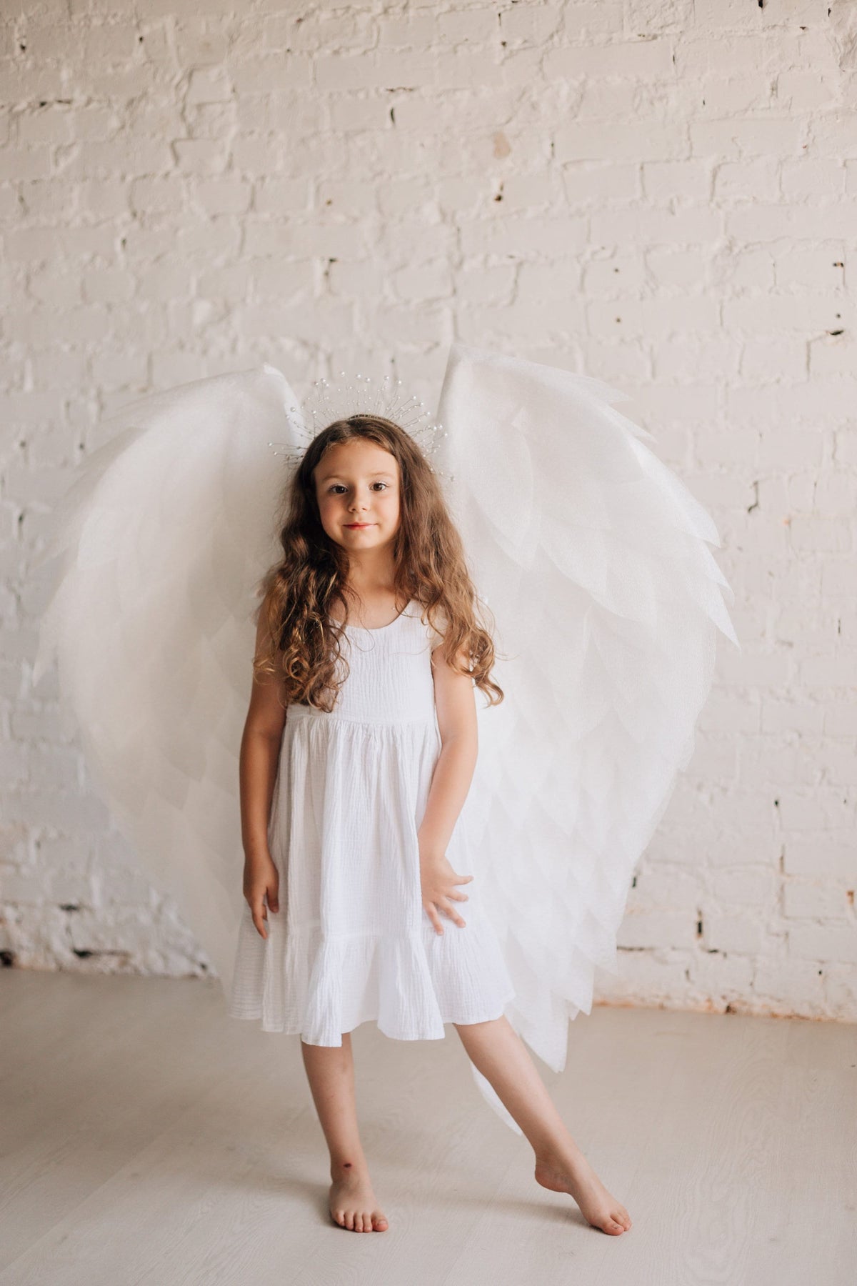 May include: A young girl wearing a white dress and angel wings with a sparkly halo. She is standing in front of a white brick wall.