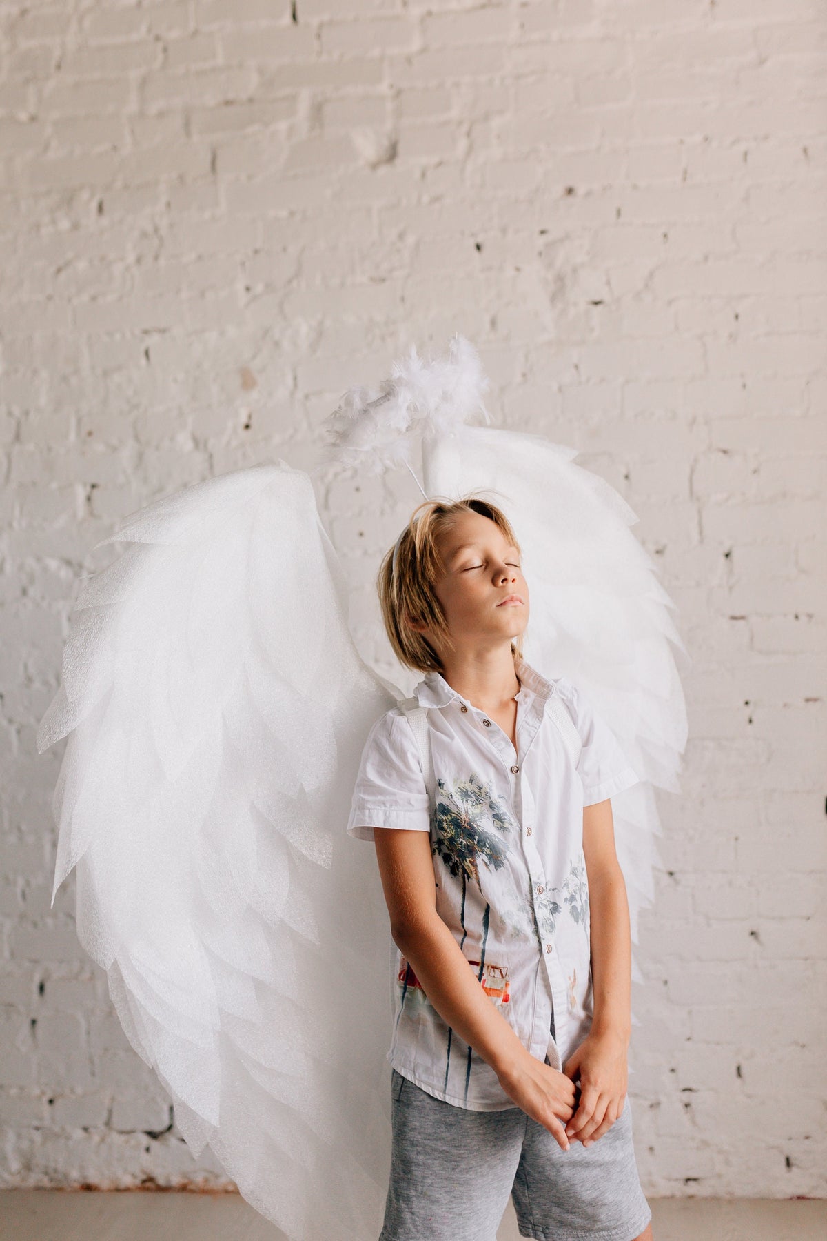 May include: A child wearing a white shirt and gray shorts, with large white angel wings and a halo. The child has their eyes closed, suggesting a serene or angelic theme. The background is a white brick wall.