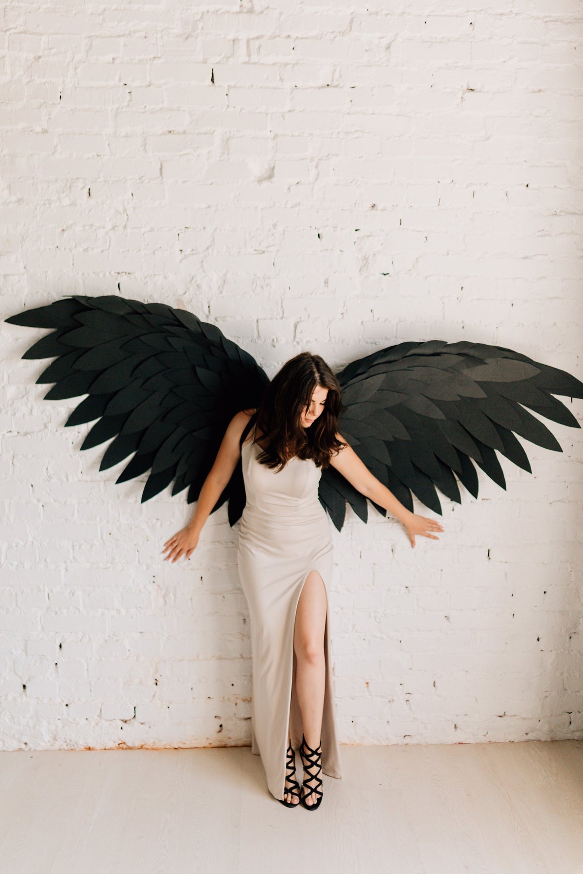 May include: A person wearing a beige dress with a high slit and black strappy heels, posing with large black paper wings against a white brick wall. The wings are detailed and spread wide.