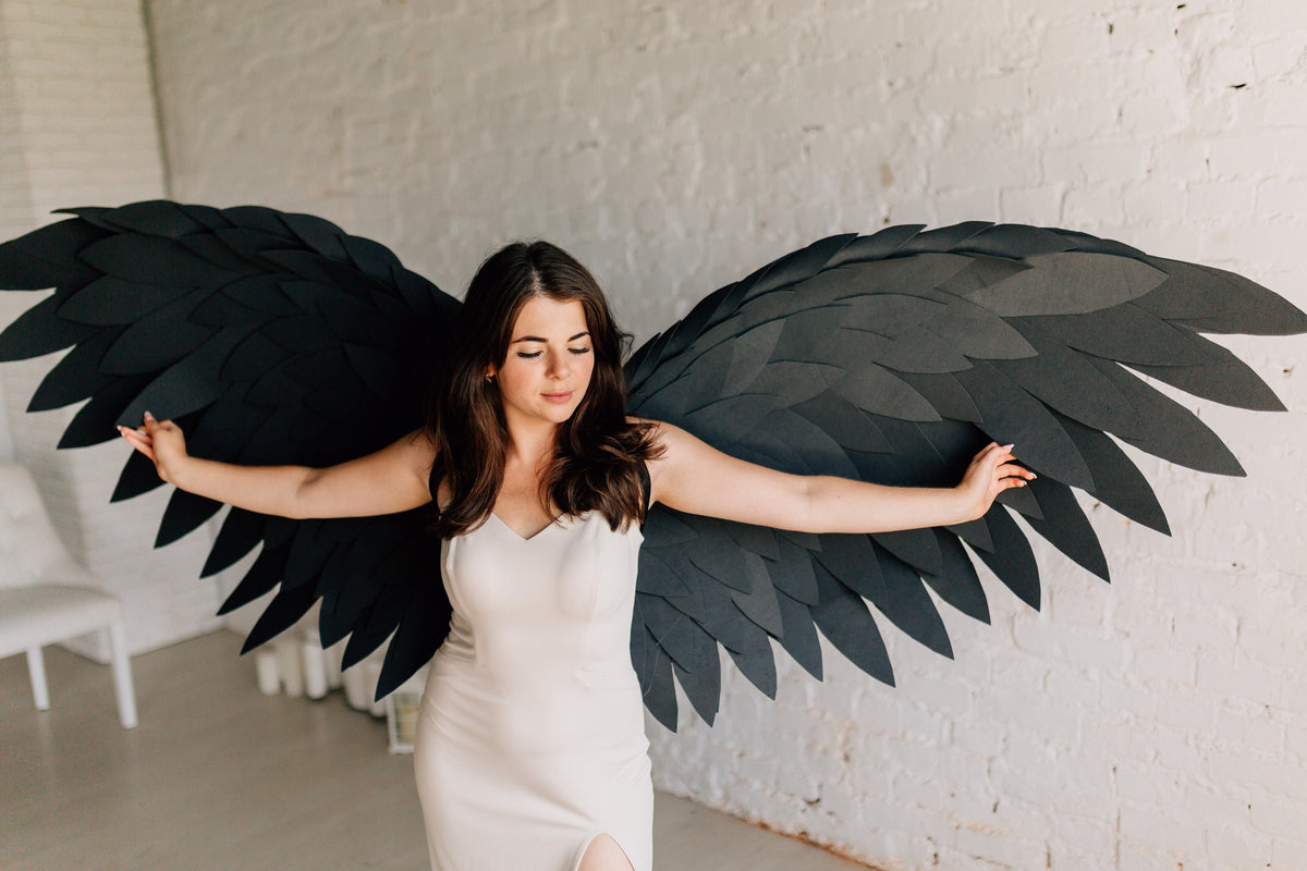 May include: A woman wearing a white dress with a high slit, posing with large, black paper wings. The wings are layered and textured, creating a dramatic effect. The background is a white brick wall.