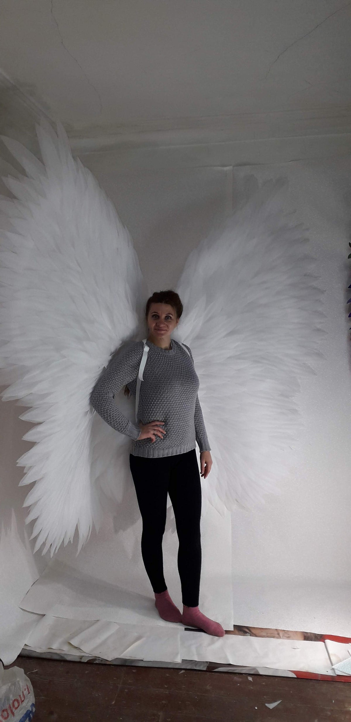 May include: A person wearing a gray sweater and black pants is standing in front of a white backdrop. They are wearing a pair of large white angel wings.