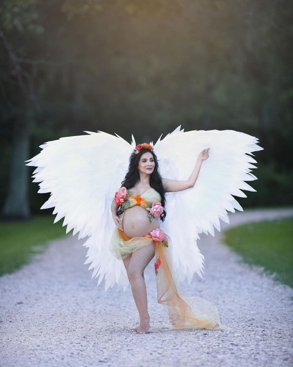 May include: A pregnant woman wearing a white angel costume with large white wings, a floral crown, and a sheer orange and yellow fabric draped around her. She is standing on a gravel path with green grass on either side.