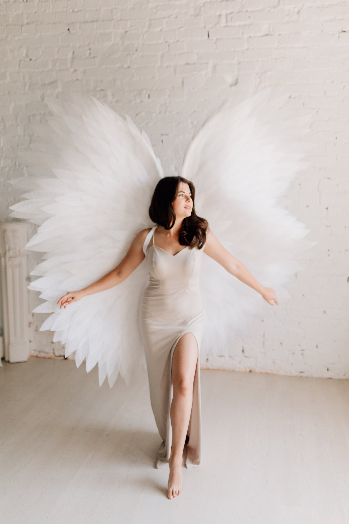 May include: A woman wearing a long, light beige dress with a slit up the leg, standing in front of a white brick wall with large white angel wings behind her.