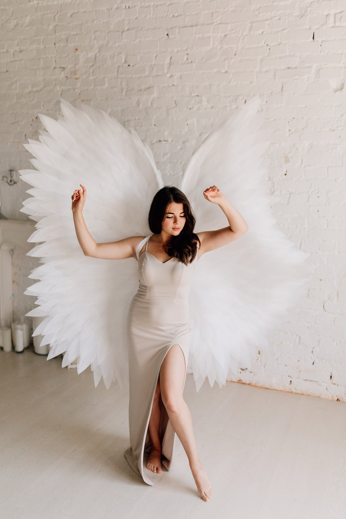 May include: A person wearing a beige dress with a slit up the leg, standing in front of a white brick wall with large white angel wings behind them.