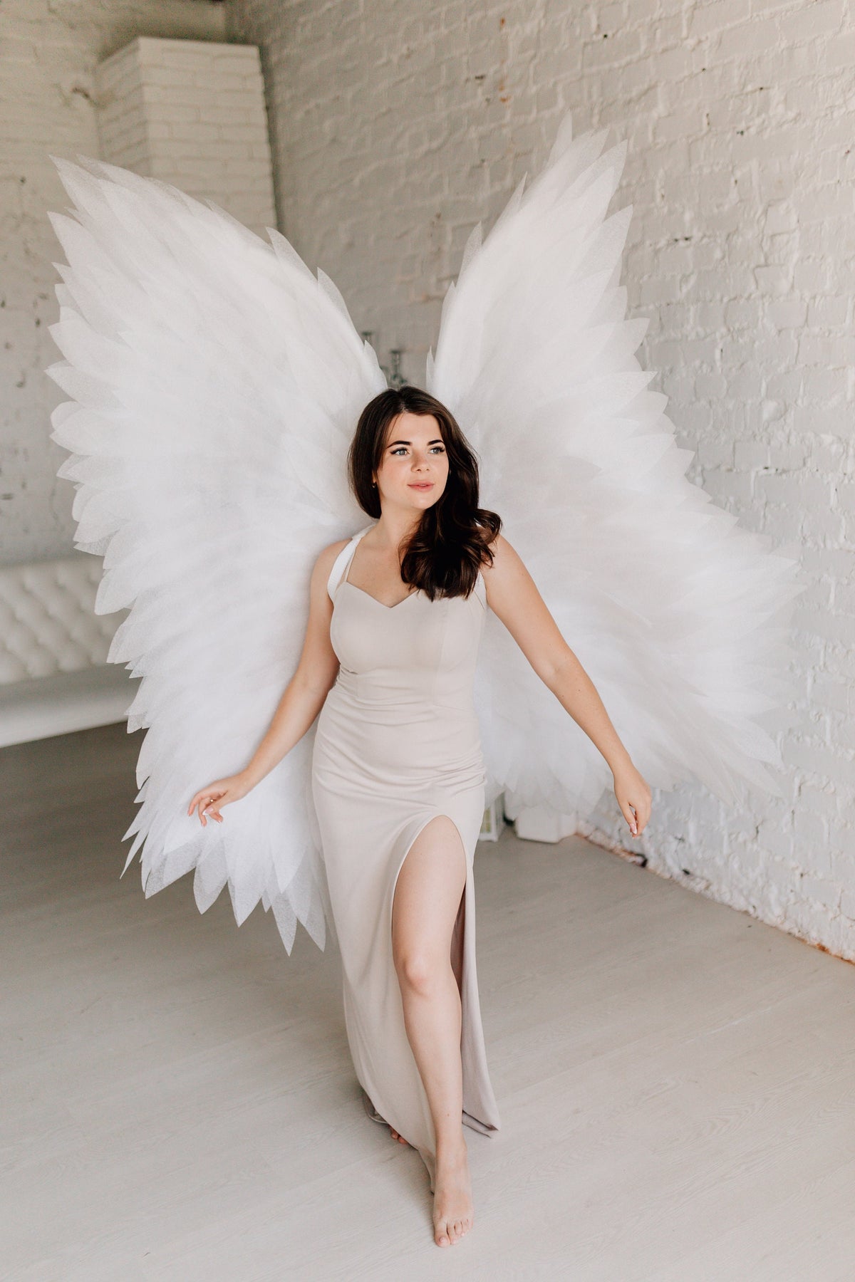 May include: A woman wearing a long, light beige dress with a slit up the leg, standing in front of a white brick wall with large white angel wings behind her.
