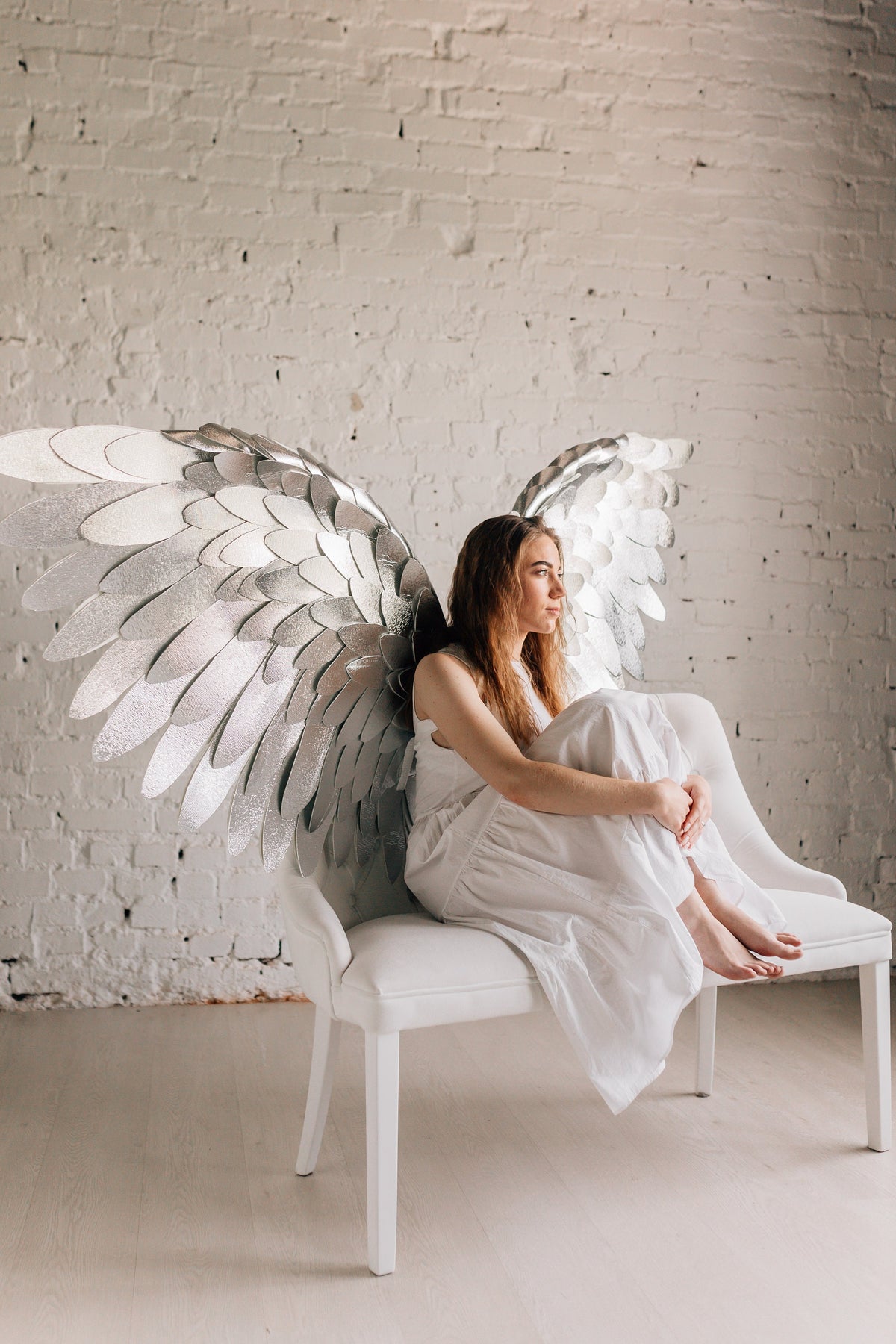 May include: A woman wearing a white dress sits on a white bench with large silver wings behind her. The wings are made of many individual feathers.