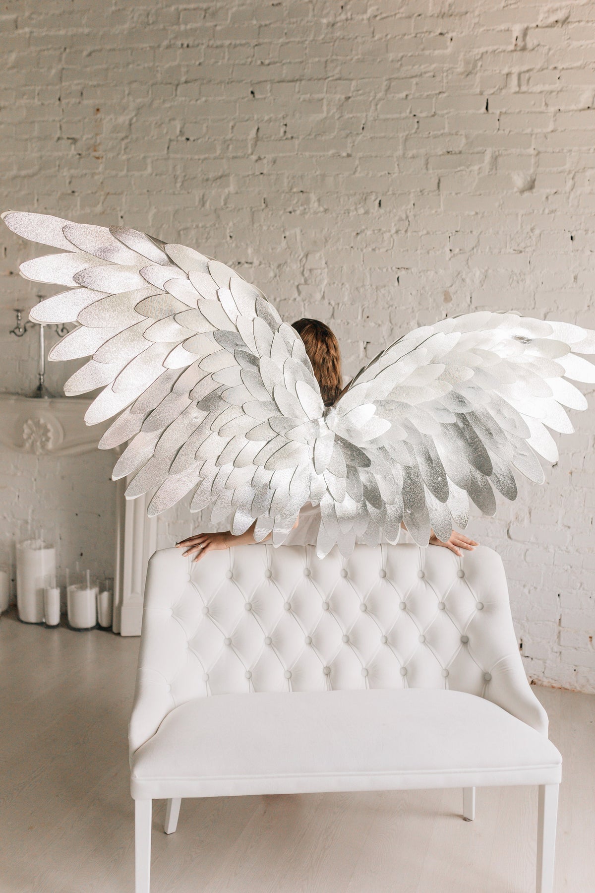 May include: A person wearing a white outfit with large silver angel wings sitting on a white tufted bench in front of a white brick wall.