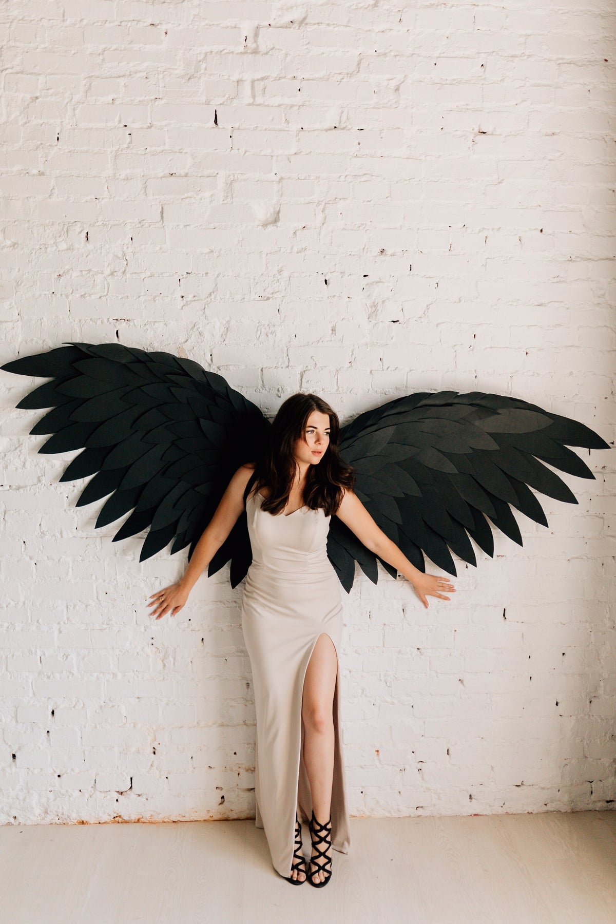 May include: A woman wearing a beige, floor-length dress with a high slit and black strappy heels, posing in front of a white brick wall. Large black paper wings are attached to the wall behind her, creating a dramatic effect.