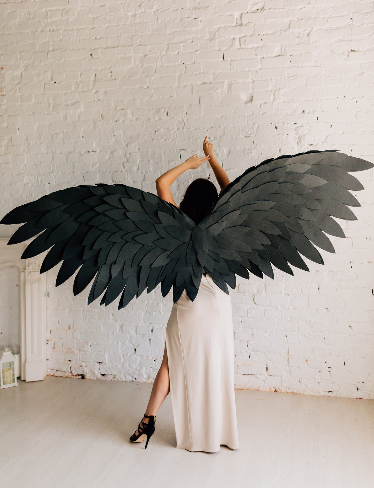 May include: A person wearing large, black paper wings, with a long, cream-colored dress and black heels. The wings are made of layered, dark gray paper, and the person's arms are raised. The background is a white brick wall.