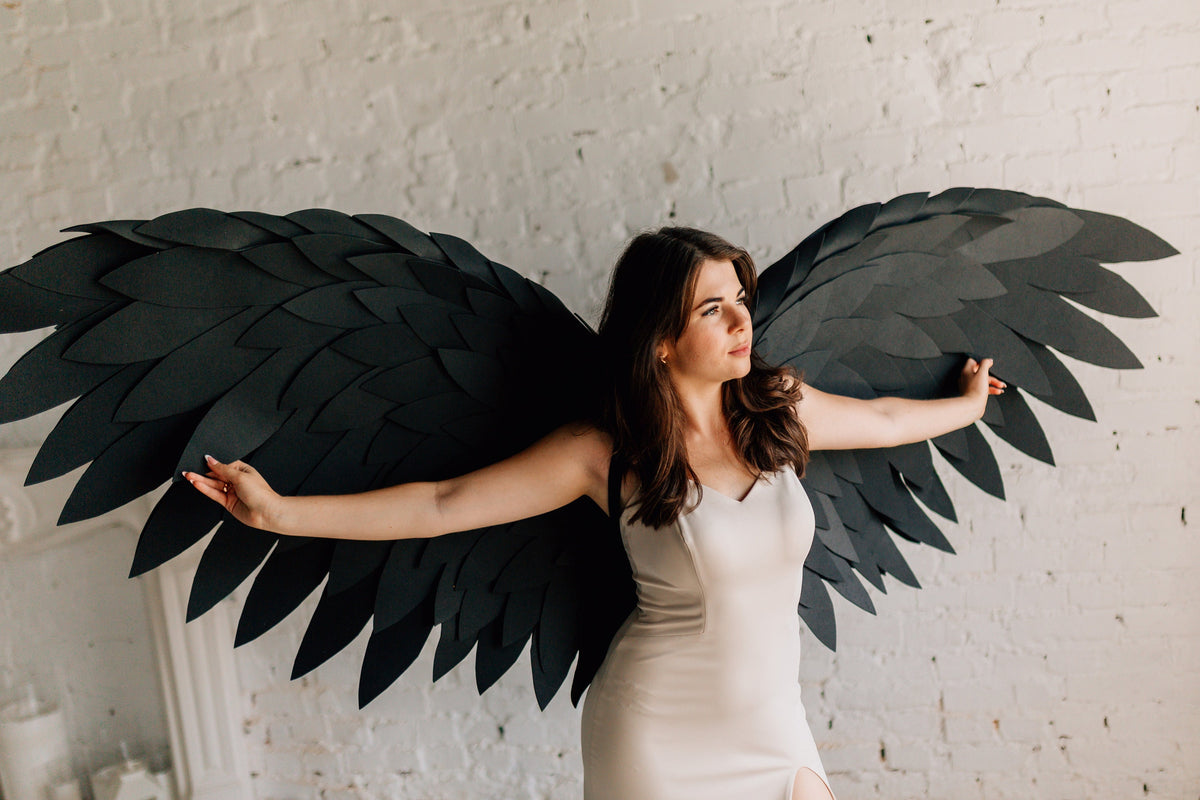 May include: Large black angel wings with layered feather design. The wings are spread wide, creating a dramatic effect. The wings are made of a dark material, possibly paper or fabric. The wings are a costume accessory.