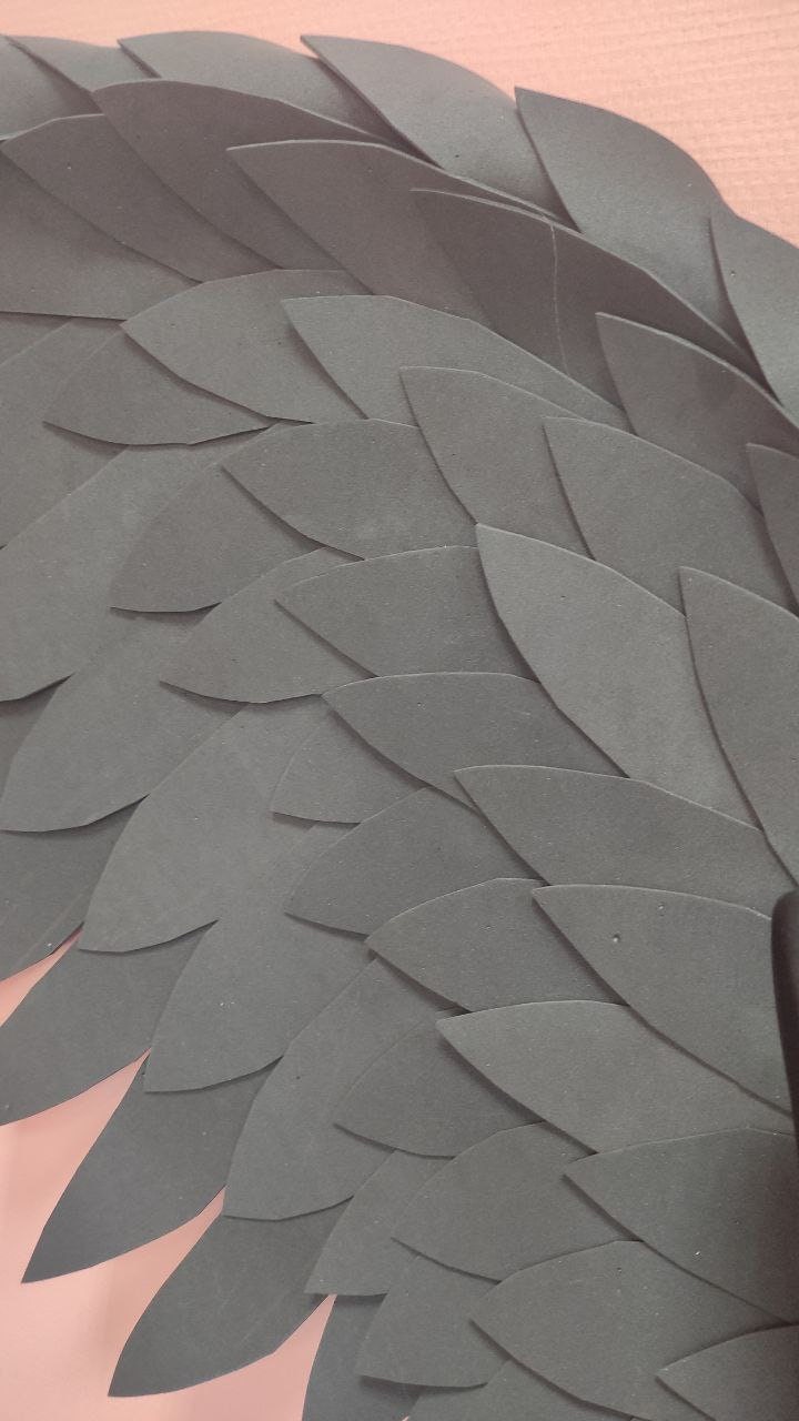 May include: Close-up of a section of gray, layered, leaf-shaped cutouts. The cutouts are arranged in an overlapping pattern, creating a textured surface. The background is a soft pink color.