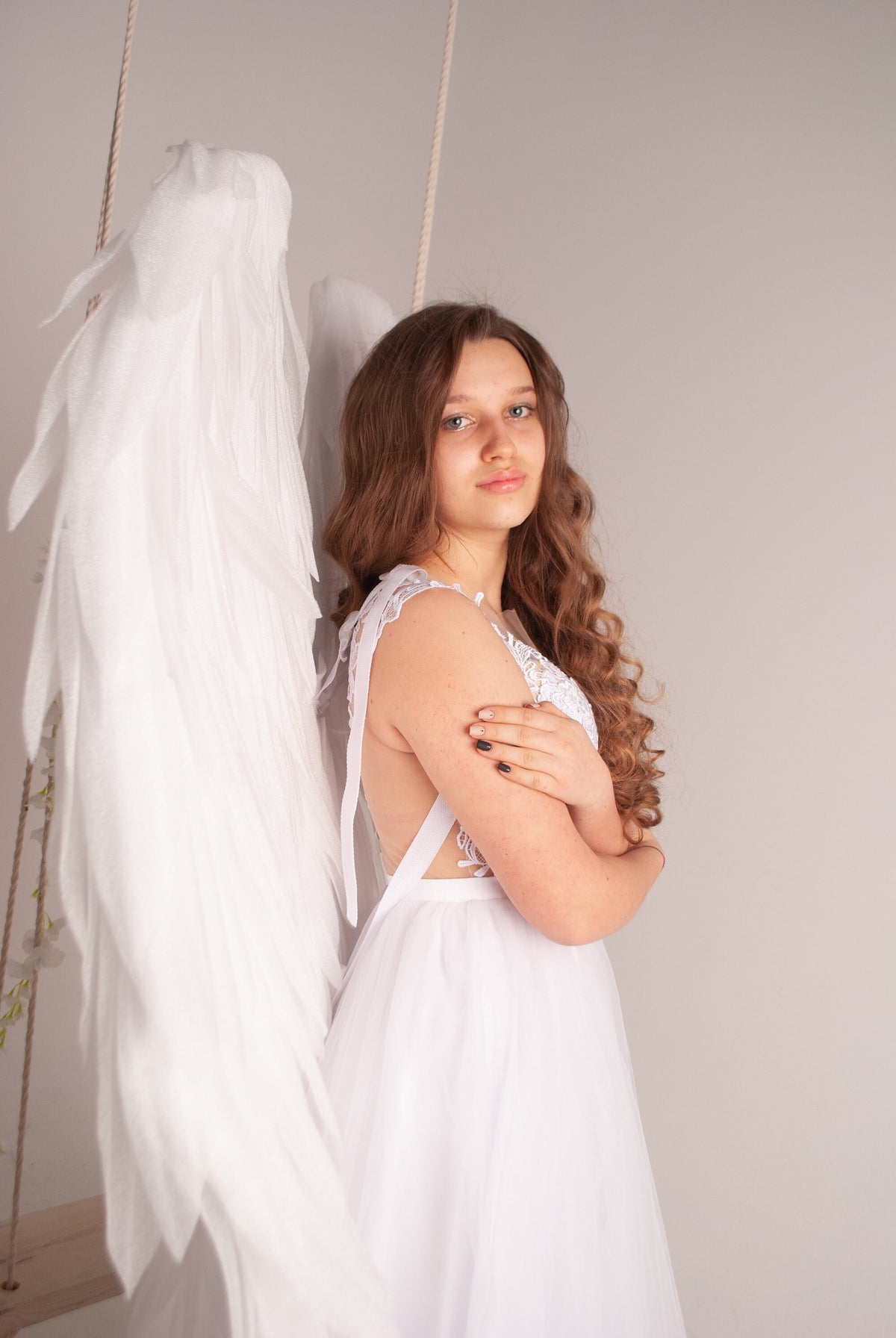 May include: A young person wearing a white dress with a tulle skirt and lace detailing. The person is posing with white angel wings attached to a swing. The background is a light gray.