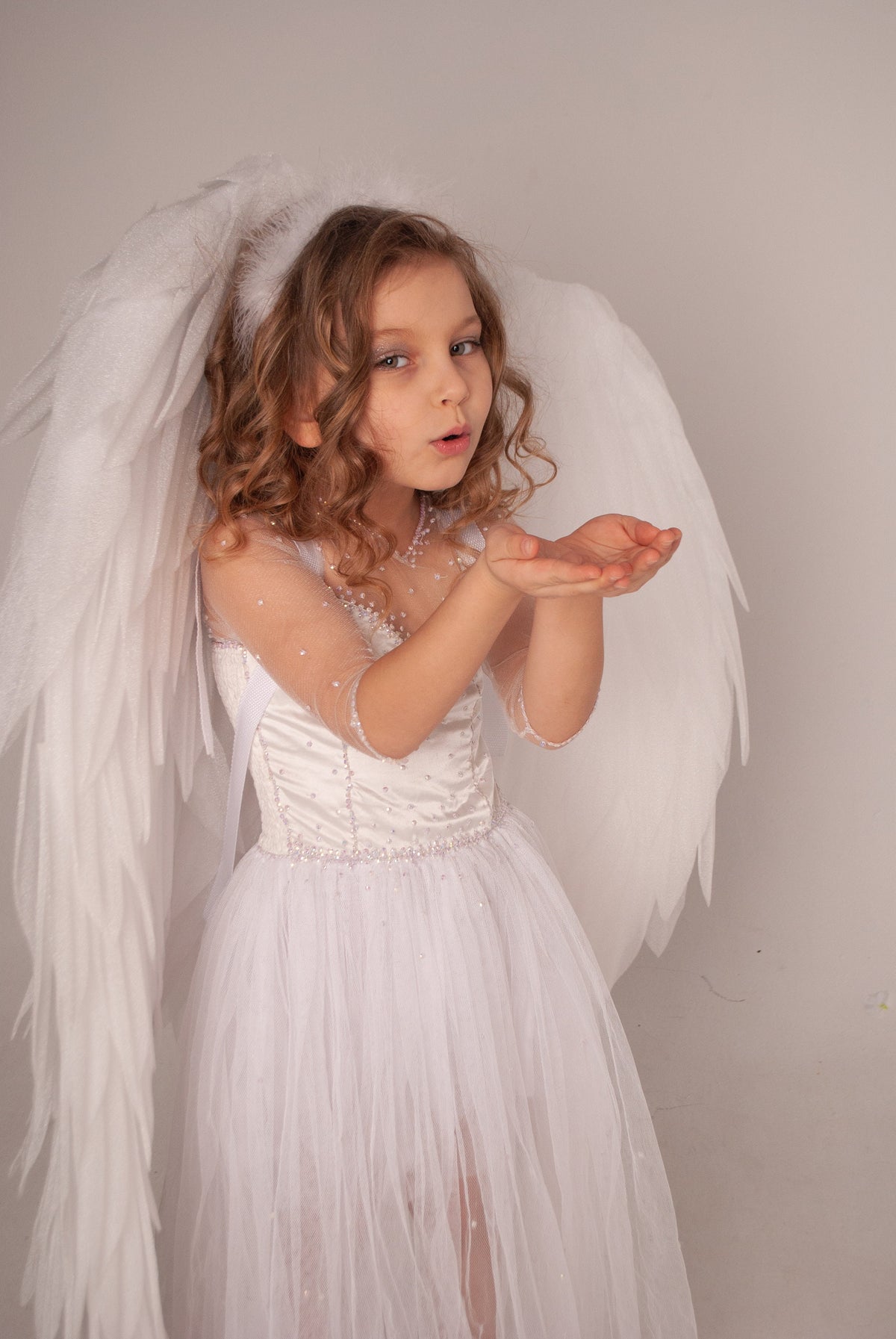 May include: A young girl dressed in a white angel costume with large feathered wings. She wears a white dress with a tulle skirt and is blowing a kiss towards the camera. The background is a plain, light gray.
