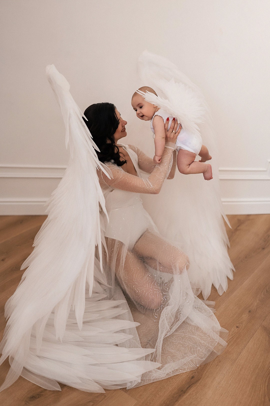 May include: A woman wearing a white dress with large white angel wings holds a baby wearing a white onesie with angel wings. The woman and baby are smiling at each other.