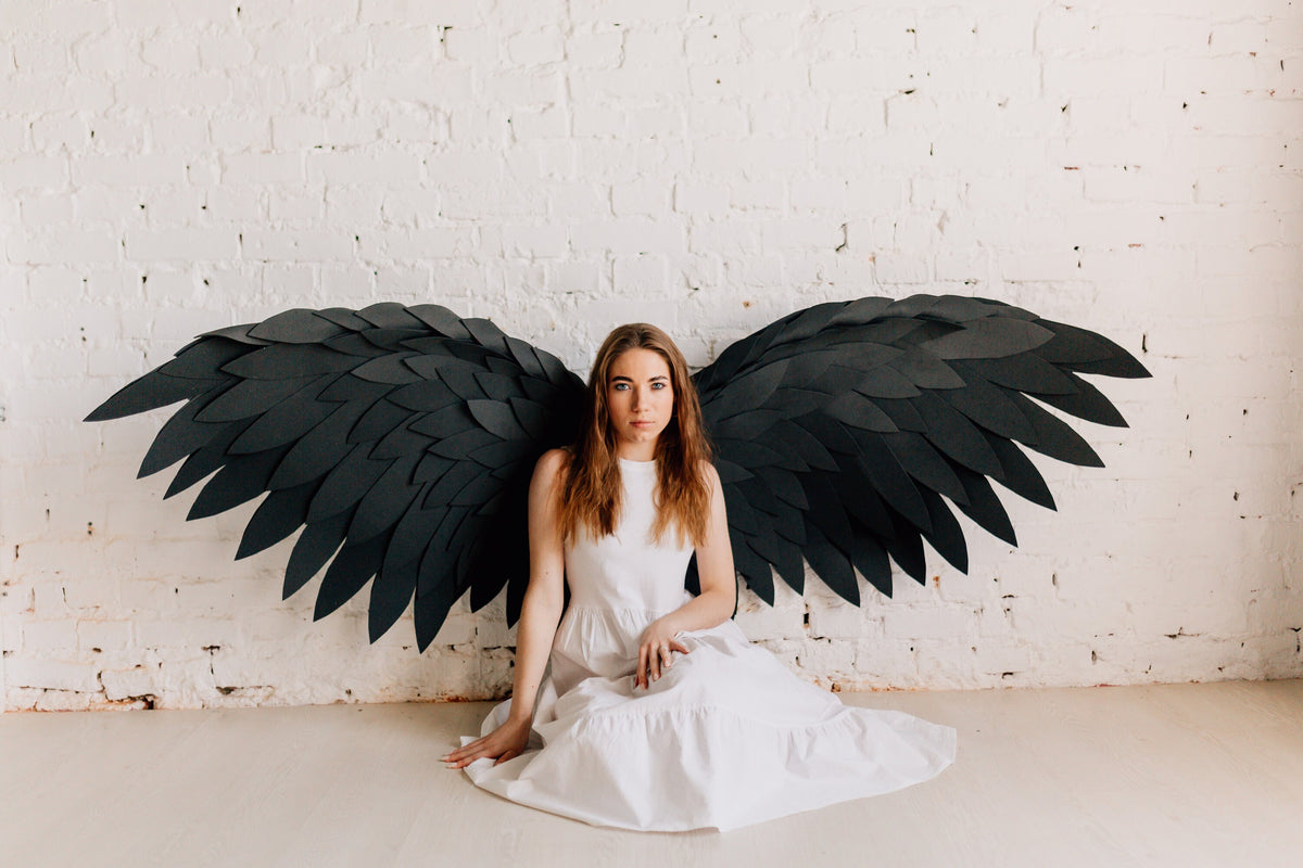 May include: A person wearing a white dress sits in front of a white brick wall with large black paper wings behind them.