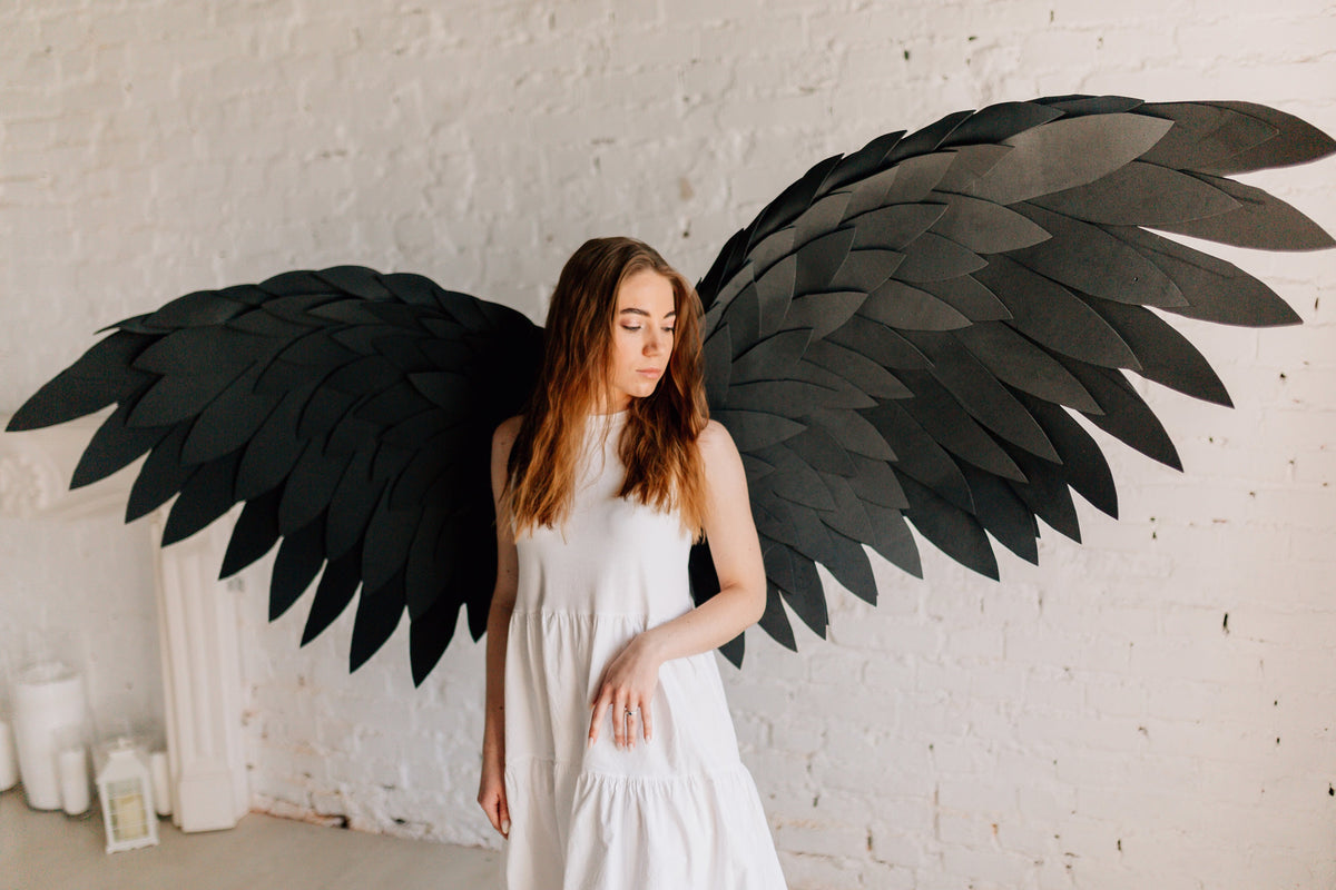 May include: A woman wearing a white dress stands in front of a white brick wall with large black wings behind her.