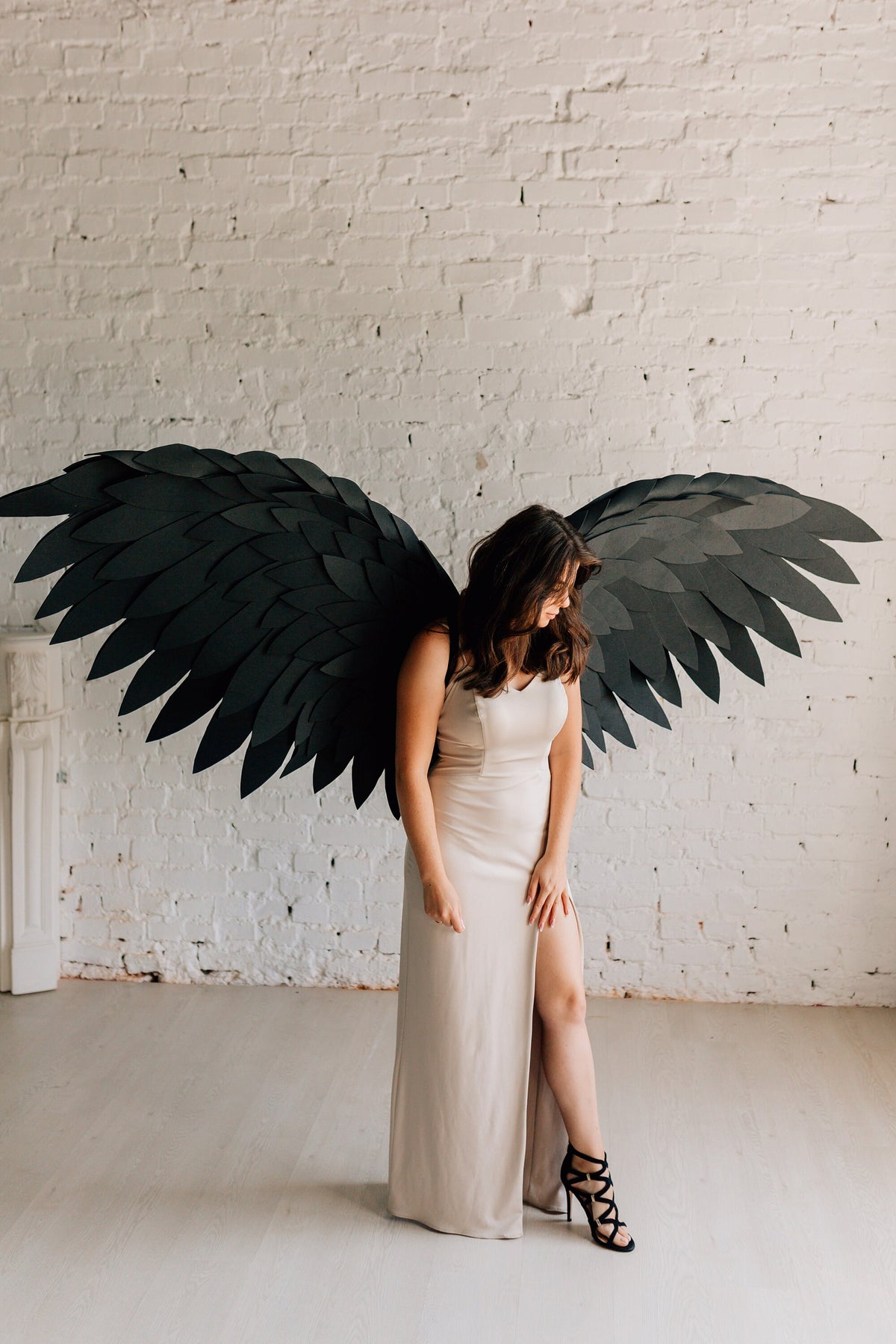 May include: A person wearing a long, light beige dress with a slit up the side, standing in front of a white brick wall. They are wearing black heels and have large black wings behind them.