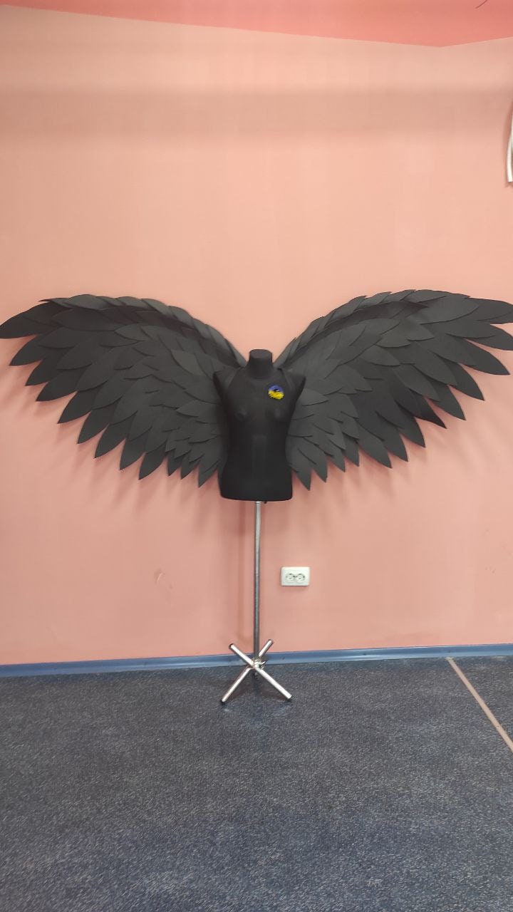May include: Black angel wings made of paper or fabric, mounted on a black mannequin on a silver stand against a pink wall.