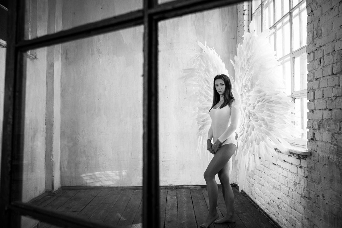 May include: A woman wearing a white bodysuit stands in a room with a large white feathered angel wing behind her. The room has a brick wall and a window with white blinds.