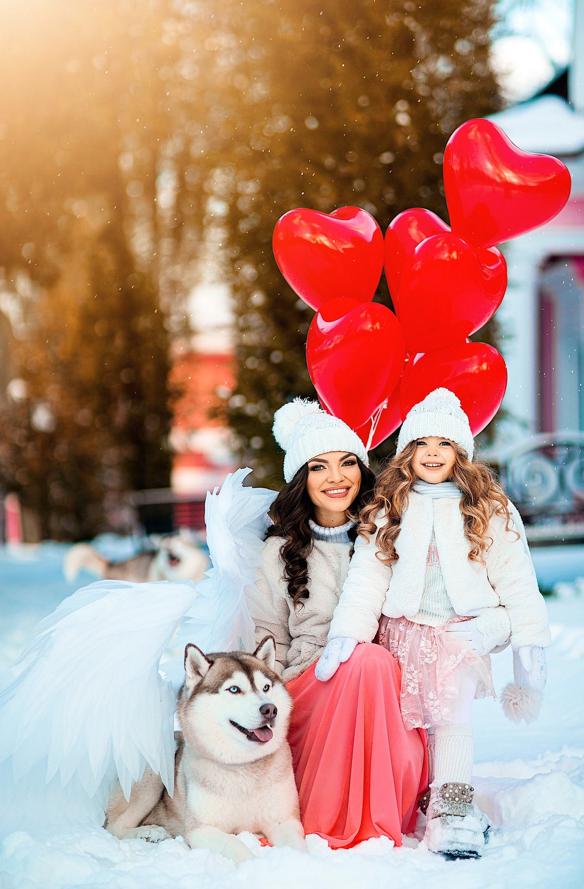 May include: Two girls, one wearing a white fur coat and a pink skirt, and the other wearing a white fur coat and a white hat, are holding red heart-shaped balloons. A husky dog is sitting in front of them. The girls are standing in a snowy winter scene.