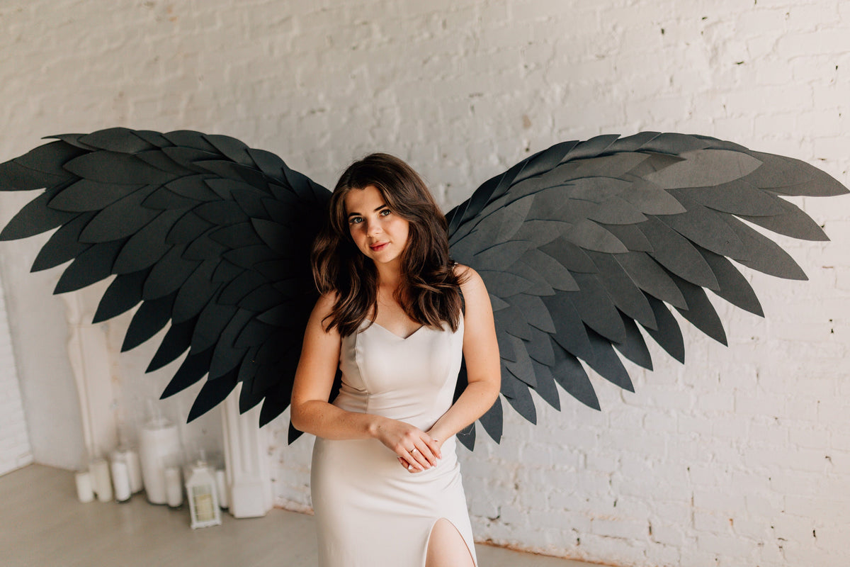 May include: A woman wearing a cream-colored dress and large black angel wings. The wings are made of layered, dark gray material, with a feather-like design. The background is a white brick wall.