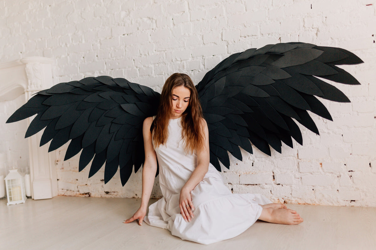 May include: Large black angel wings with layered feather design. The wings are displayed against a white brick wall. The person is wearing a white dress and is sitting on the floor.