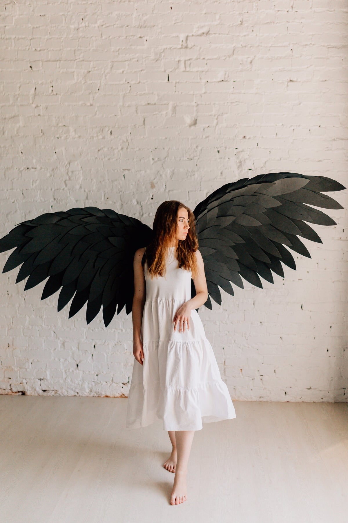 May include: A woman wearing a white tiered dress and large black paper wings stands in front of a white brick wall. The wings are detailed with layered, feather-like cutouts. The woman is barefoot.