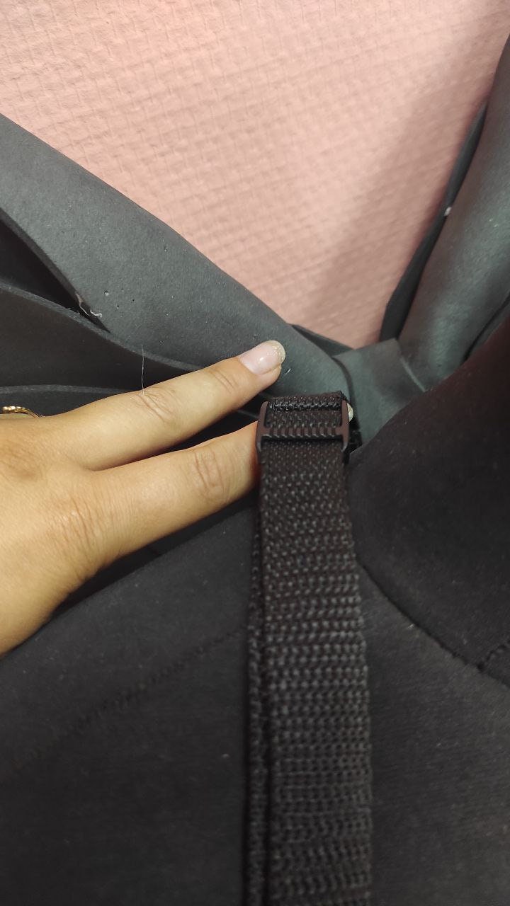 May include: Close-up of a black strap with a plastic buckle, likely part of a bag or backpack. The strap is made of a woven material. The background is a soft pink.