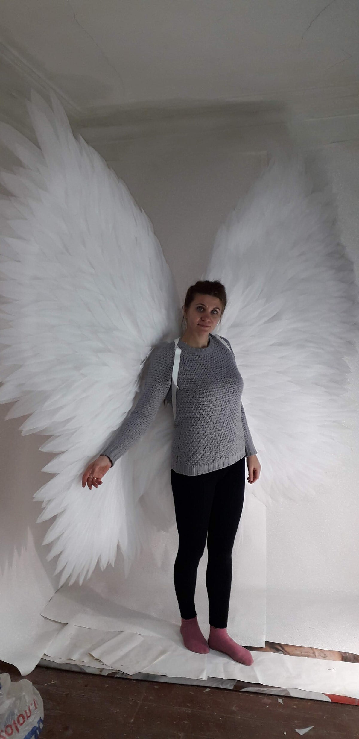May include: A person wearing a gray sweater and black pants is standing in front of a white backdrop. They are wearing a pair of large, white angel wings.