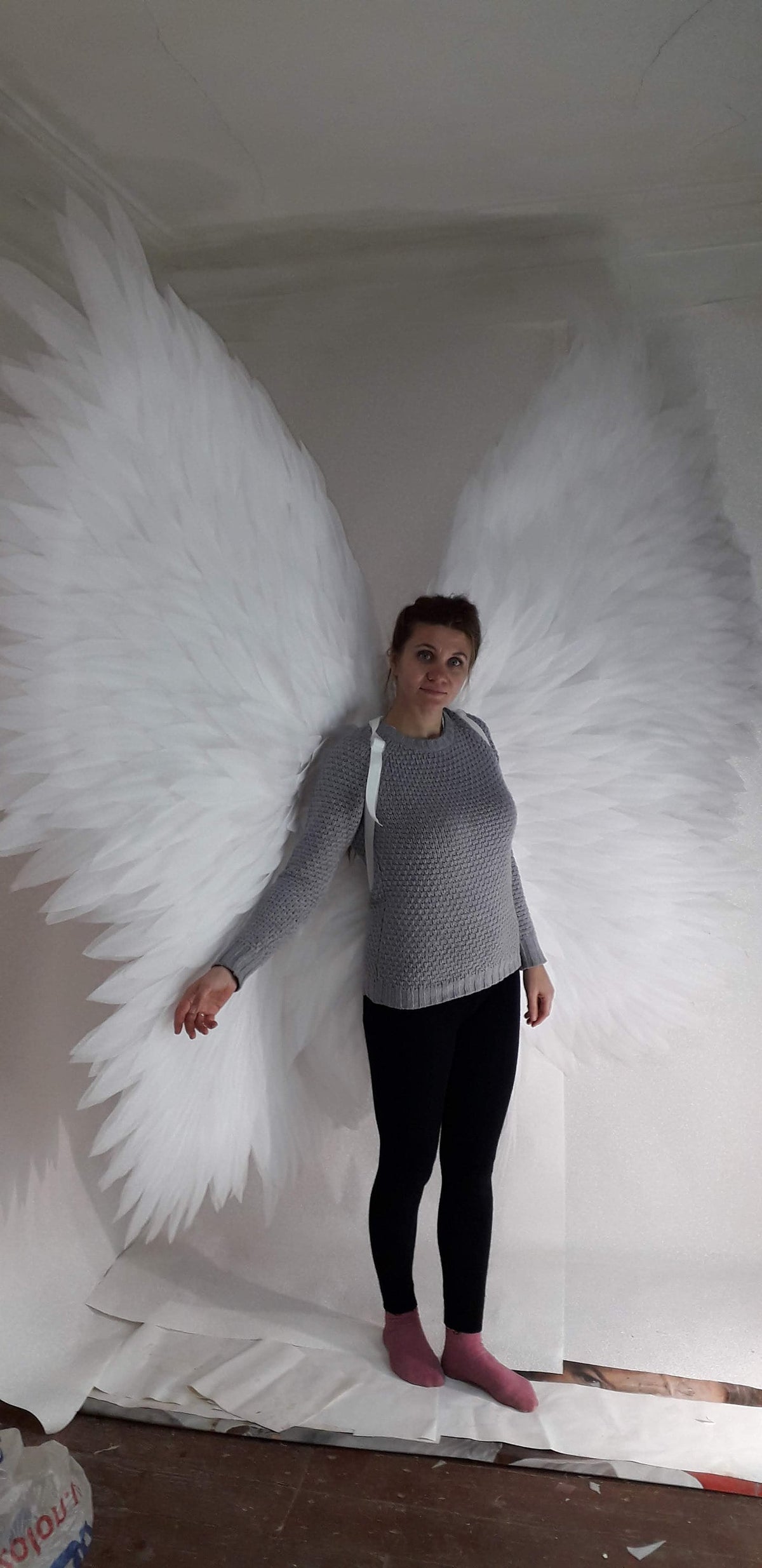 May include: A person wearing a gray sweater and black pants is standing in front of a white backdrop. They are wearing a pair of large, white angel wings.