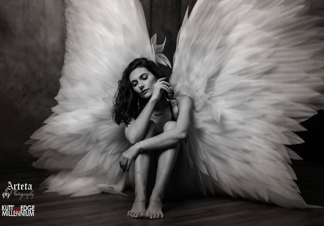 May include: A woman with long dark hair sits with her legs crossed, wearing a black top and white angel wings. She is looking down and holding her hand to her chin. The background is a dark gray wall.