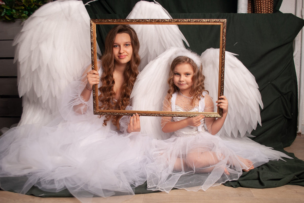 May include: Two individuals dressed in white angel costumes with large feathered wings pose with a gold-framed rectangle. The individuals are seated, surrounded by white tulle, and have halos. The background includes a dark green fabric and a wooden wall.