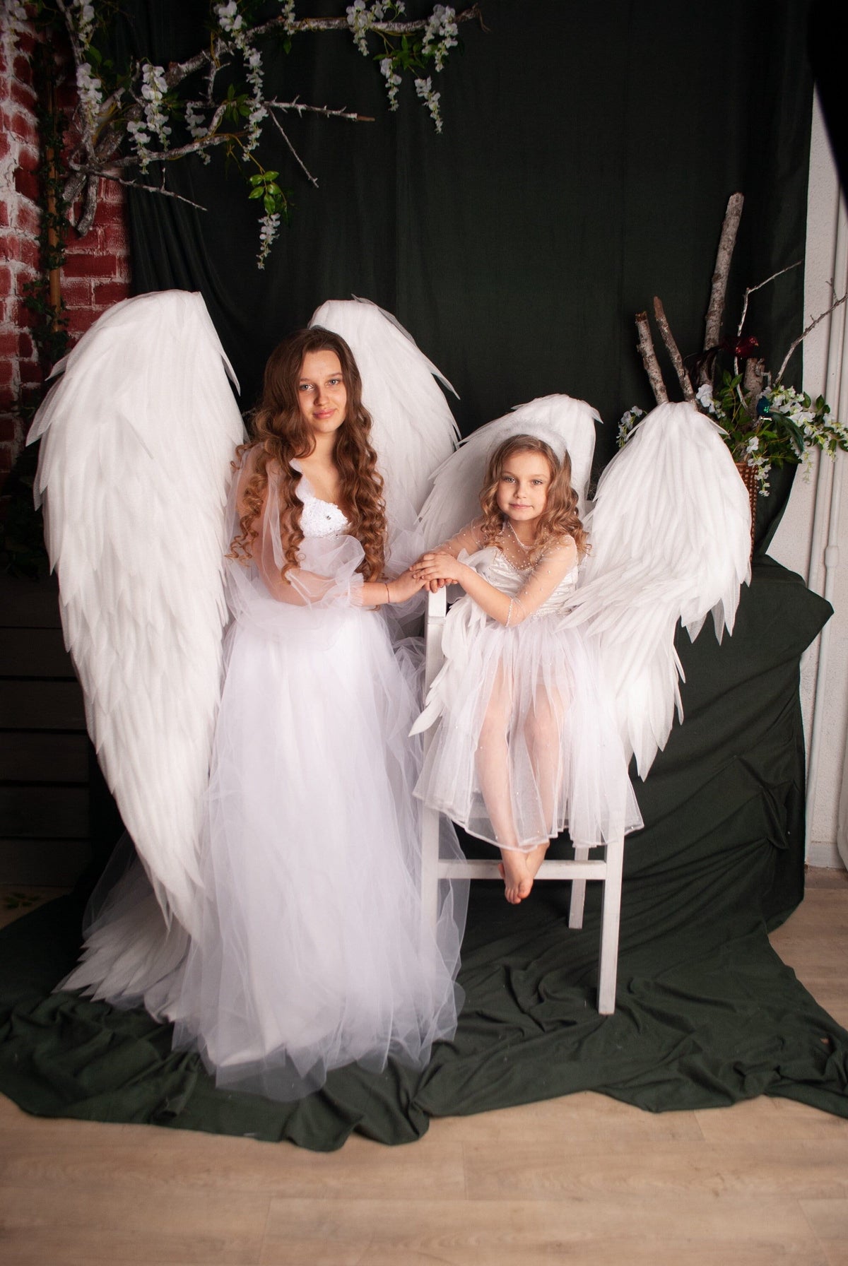 May include: Two individuals wearing white angel wings and dresses pose for a photo. One stands, the other sits on a white chair. The wings are large and feathered. The background is a dark green curtain and a brick wall.