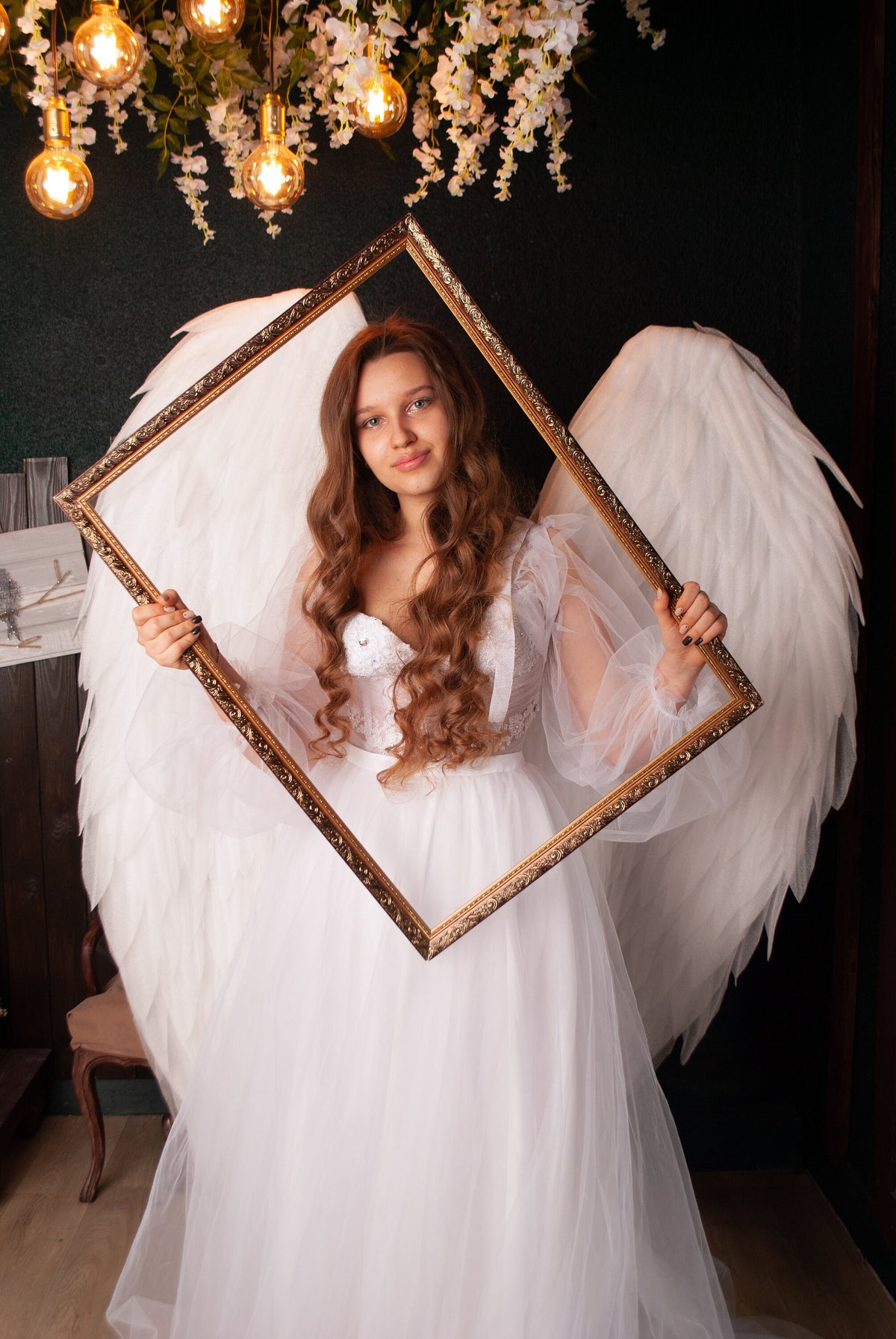 May include: A woman in a white dress and large white feathered wings holds a gold-framed square. The woman has long, wavy auburn hair. The background is dark with decorative lights and floral accents.