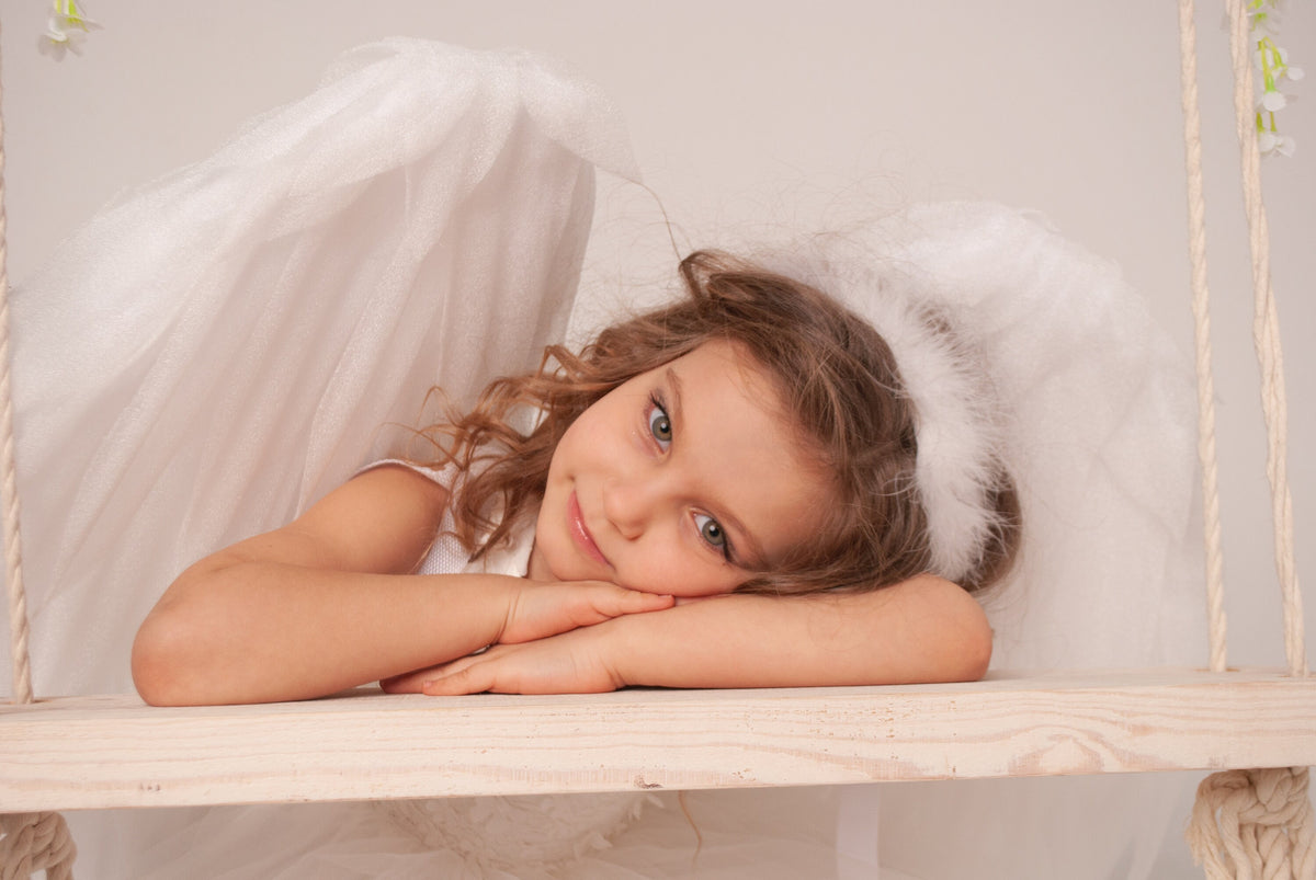 May include: A young person with curly brown hair wearing a white dress, angel wings, and a halo, resting on a wooden swing. The background is a soft, neutral color, creating a serene and angelic atmosphere.