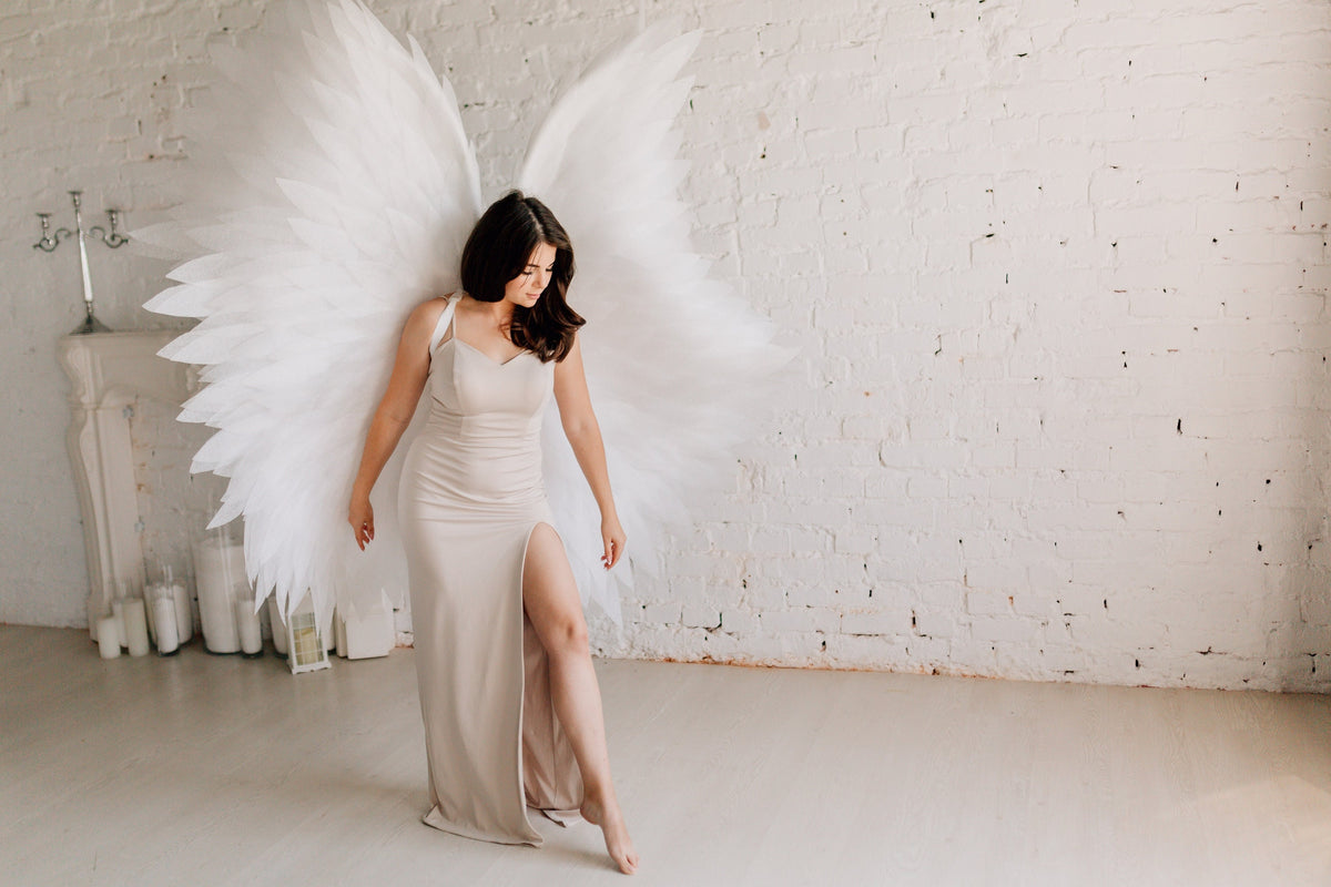 May include: A woman wearing a long, light brown dress with a slit up the side, standing in front of a white brick wall. She is wearing large, white feathered angel wings.